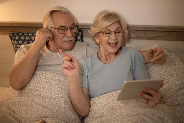 Gelukkig senior vrouw ondertekenen tijdens het luisteren naar muziek met haar man op touchpad in slaapkamer