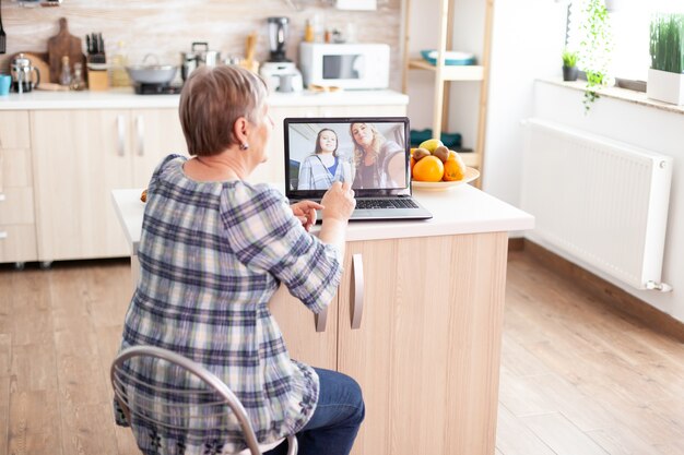 Gelukkig senior vrouw tijdens een videoconferentie met familie met behulp van laptop in de keuken. Online bellen met dochter en nichtje. Oude bejaarde die moderne communicatie online internet webtechnologie gebruikt.