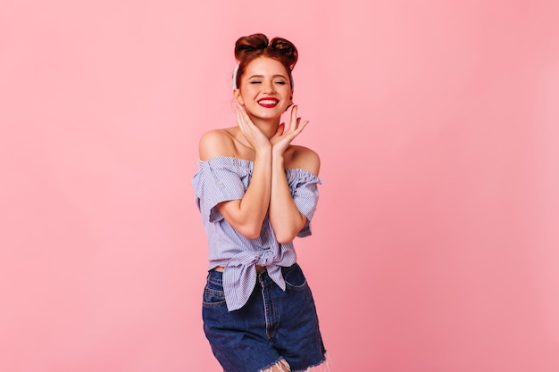 Gelukkig stijlvol meisje met pinup kapsel staande op roze achtergrond Aantrekkelijke gember jonge vrouw die positieve emoties uitdrukt