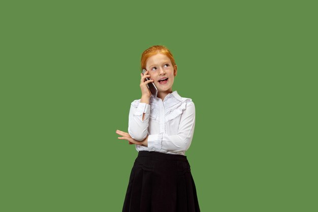 Gelukkig tiener meisje permanent, glimlachend met mobiele telefoon over trendy groene studio achtergrond. Mooi vrouwelijk portret van halve lengte. Menselijke emoties, gezichtsuitdrukking concept.