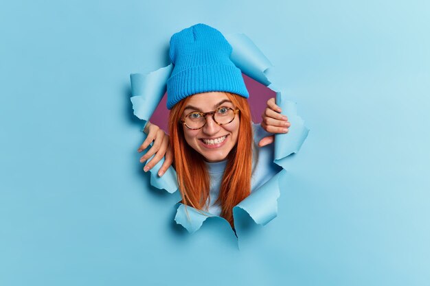 Gelukkig tienermeisje breekt door papieren muur heeft plezier blikken met plezier draagt blauwe hoed bril glimlacht toothily