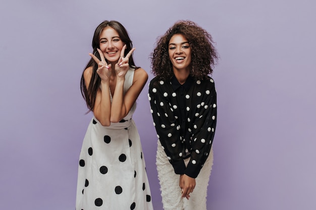 Gelukkig trendy meisjes met donkerbruin kapsel in polka dot zwart-witte kleren die camera onderzoeken en op geïsoleerde muur glimlachen