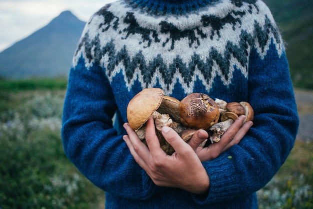 Gelukkig, trotse picker man in traditionele blauwe wollen trui met ornamenten staat op camping in de bergen, houdt stapel van heerlijke en biologische paddestoelen in de armen