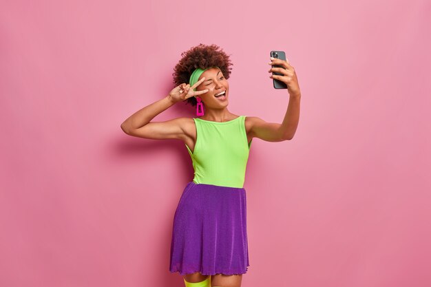 Gelukkig uitgaande vrouw maakt overwinning gebaar, oog knipoogt en vormt op de camera van de smartphone, neemt selfie, gekleed in kleurrijke outfit