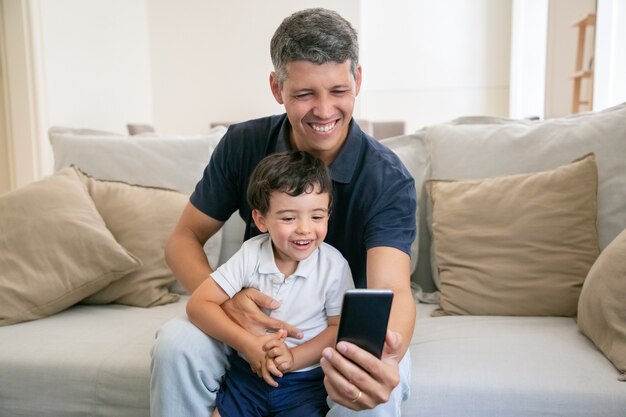 Gelukkig vader en schattig zoontje plezier samen, met behulp van telefoon voor videochat zittend op de bank thuis