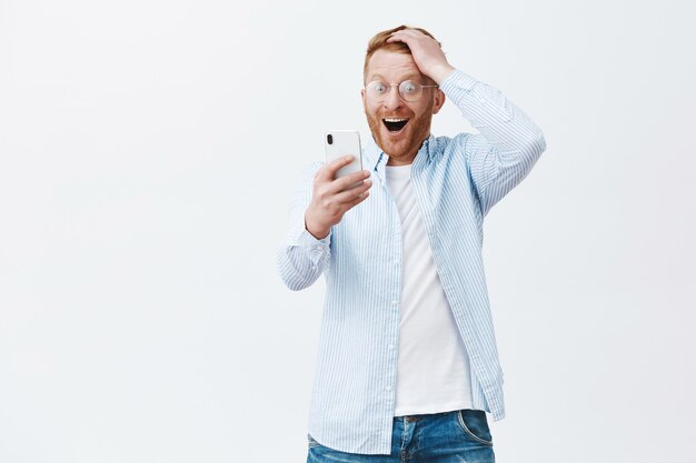 Gelukkig verheugende Europese man in bril en shirt, kapsel aan te raken terwijl hij tevreden en verbaasd naar het smartphonescherm staarde