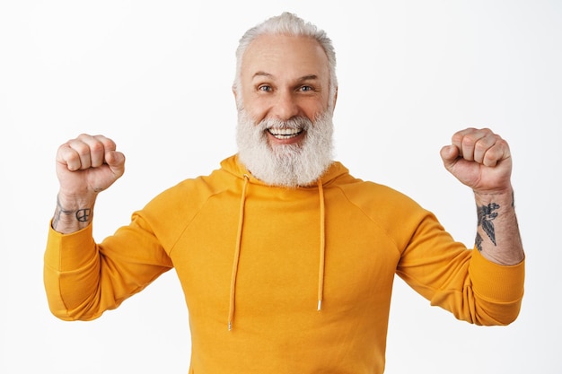 Gelukkig volwassen man met baard en tatoeages vieren van succes en overwinning, balde vuisten en schud ze in triomf, doel bereiken, winnen en lachen met vreugde, staande op witte achtergrond