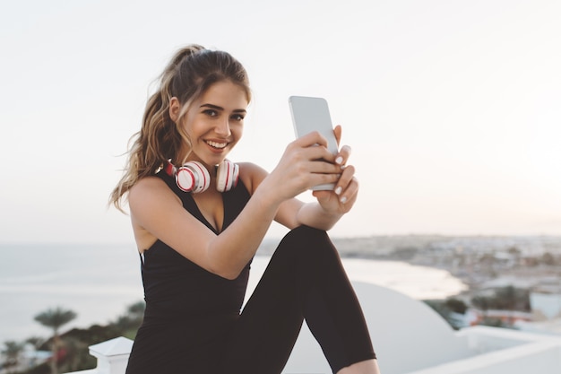 Gelukkig vreugdevolle jonge vrouw in aantrekkelijke sportkleding selfie maken op telefoon, glimlachen, genieten van zonsopgang in de ochtend aan de kust. Vrolijke stemming, echt geluk