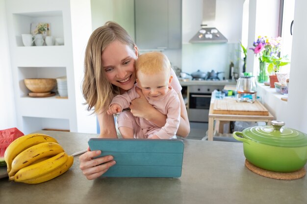 Gelukkig vreugdevolle moeder en baby dochter kijken naar online recepten, met behulp van tablet in de keuken, samen glimlachen op scherm. Kinderopvang of koken thuis concept