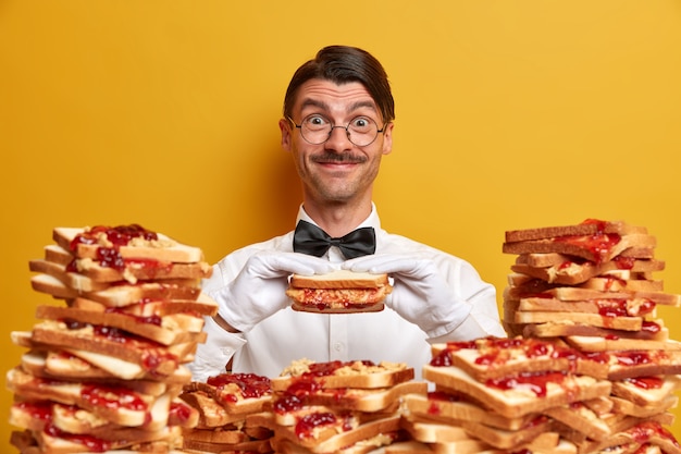 Gelukkig vriendelijke ober heeft gelegenheid om heerlijke sandwich te proeven, poseert in de buurt van stapel brood toast, draagt formele outfit en witte handschoenen, geïsoleerd op gele muur. Tijd om hamburgers te eten