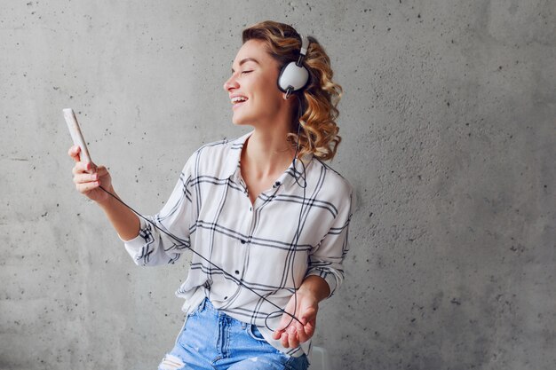 Gelukkig vrij blonde hipster vrouw genieten van muziek via oortelefoons, zittend op een stoel op grijze stedelijke muur achtergrond.