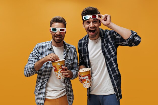 Gelukkig vrolijke brunet mannen in witte t-shirts en blauw geruite shirts glimlach pose in 3d-brillen eten popcorn en kijken naar film op oranje achtergrond