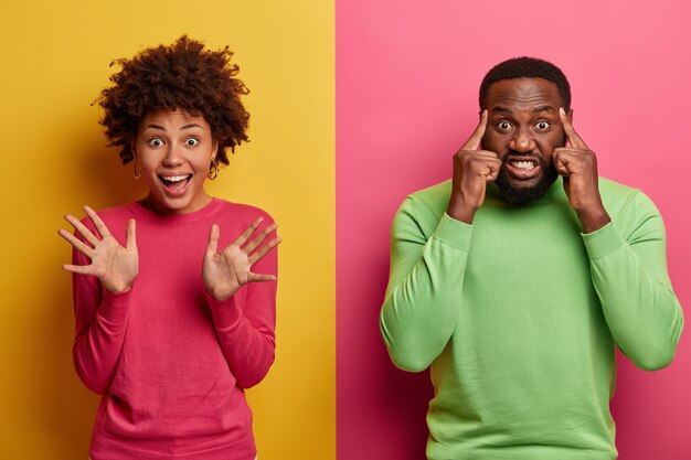 Gelukkig vrolijke vrouw heft handpalmen op, zwaait met de handen hallo, maakt vriendelijk gebaar, intense bebaarde man raakt tempels, probeert zich te concentreren, klemt tanden, heeft hoofdpijn, gaat tegen twee gekleurde muur staan