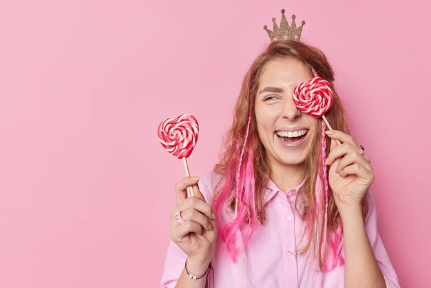 Gelukkig vrolijke vrouw met lang haar bedekt ogen met zoete snoepjes op stokken heeft vrolijke stemming glimlacht breed draagt kleine kroon en shirt geïsoleerd over roze achtergrond lege kopie ruimte aan de linkerkant