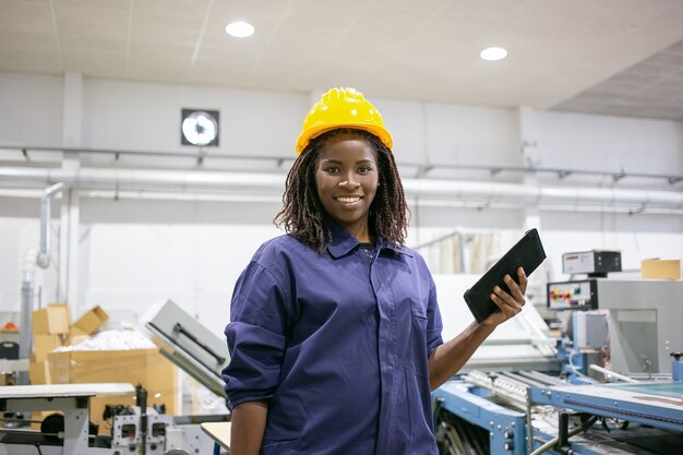 Gelukkig vrouwelijke fabrieksarbeider in veiligheidshelm en over het algemeen, staande op de machine, tablet vasthouden, frontaal kijken en glimlachen