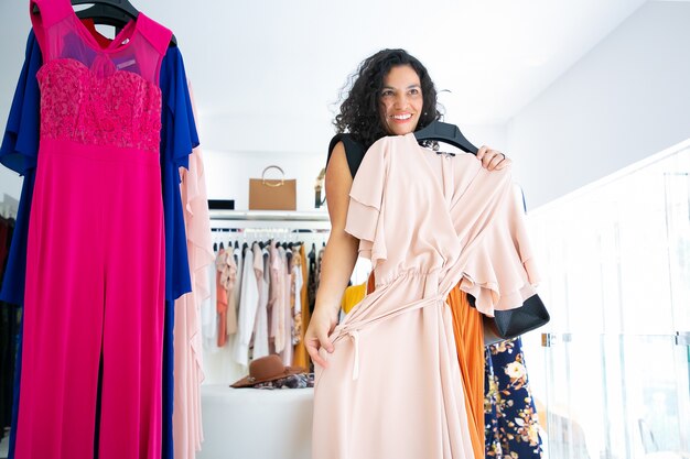 Gelukkig vrouwelijke klant jurk met hanger toe te passen en in spiegel kijken. Vrouw die kleren in modewinkel kiest. Winkelen of winkelconcept