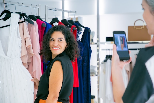 Gelukkig vrouwelijke shoppers genieten van winkelen in kledingwinkel samen, jurk aanraken, poseren en fotograferen op mobiele telefoon. Consumentisme of winkelconcept