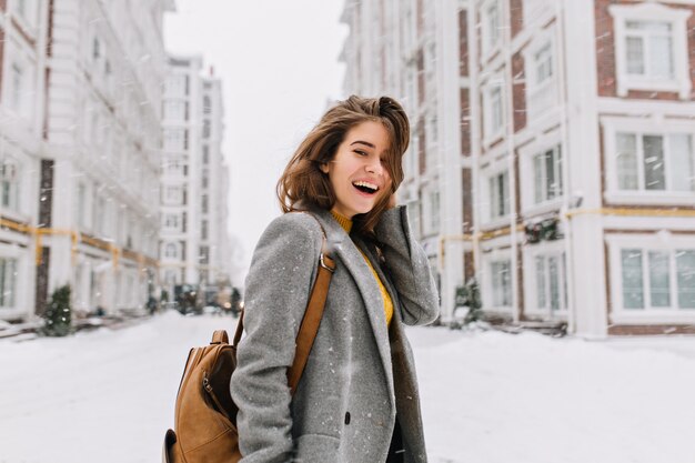 Gelukkig wintertijd in grote stad van charmante vrouw lopen op straat in jas met rugzak. Genieten van sneeuwval, positiviteit uiten, glimlachen, vrolijke, opgewekte stemming, ware emoties, nieuwjaarsstemming.