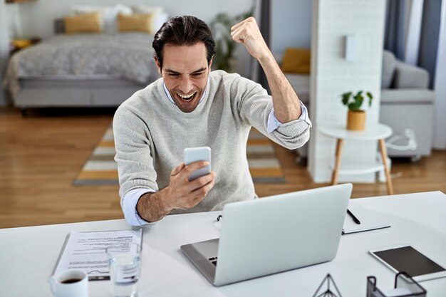 Gelukkig zakenman vieren tijdens het lezen van goed nieuws op mobiele telefoon thuis