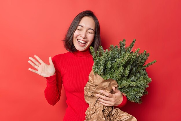 Gelukkig zorgeloze jonge Aziatische vrouw heeft feestelijke stemming giechelt positief sluit ogen draagt coltrui houdt groen sparren boeket gewikkeld in papier poses tegen rode achtergrond. Nieuwjaarsvoorbereiding