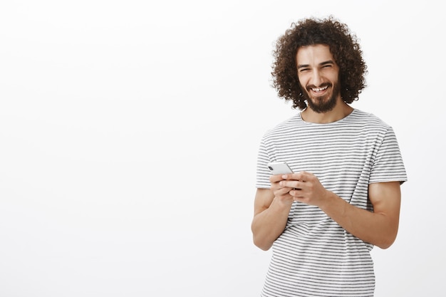 Gratis foto gelukkig zorgeloze knappe man met baard en afro kapsel, smartphone vast te houden en te lachen