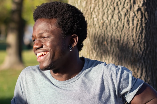 Gelukkig zwarte man tijd buitenshuis doorbrengen in stadspark op zonnige dag. Aantrekkelijke Afro-Amerikaanse man in grijs T-shirt die opzij kijkt en vrolijk lacht. Close-up shot. Jeugd, levensstijl, houding concept.