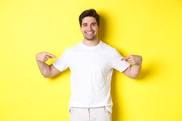 Gelukkige aantrekkelijke man die met de vingers naar je logo wijst, promo op zijn t-shirt laat zien, staande op een gele achtergrond.