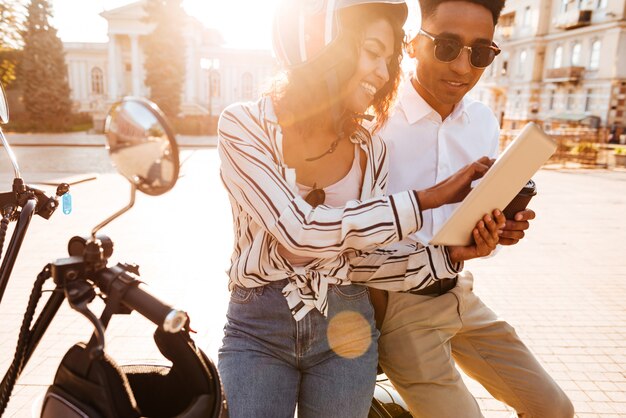 Gelukkige afrikaanse paar zittend op moderne motor en met behulp van tablet-computer op straat