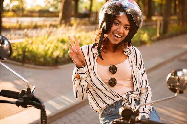 Gratis foto gelukkige afrikaanse vrouw in de zitting van de motohelm op moderne motor in openlucht terwijl het bekijken en het tonen van vrede bij de camera
