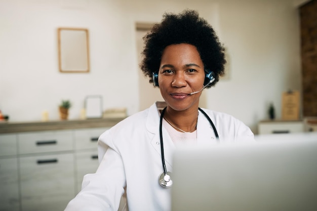 Gelukkige Afro-Amerikaanse arts met hoofdtelefoon die op laptop op haar kantoor werkt