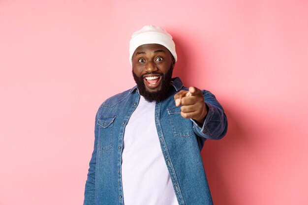 Gelukkige Afro-Amerikaanse hipster man wijzende vinger naar camera, heb je nodig, glimlachend opgewonden, staande over roze achtergrond