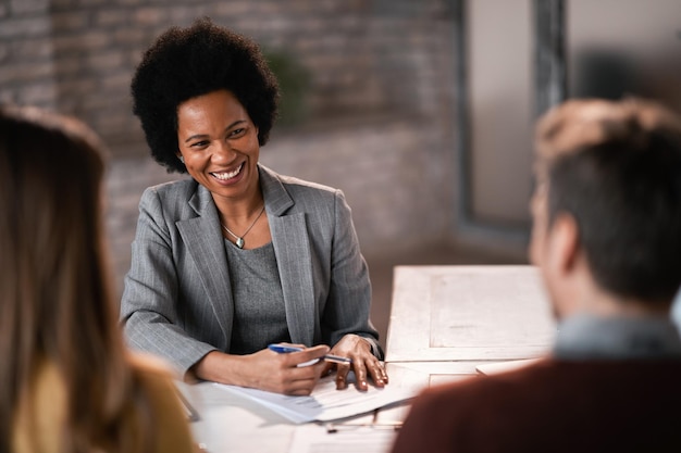 Gelukkige Afro-Amerikaanse makelaar die aan papierwerk werkt terwijl hij een ontmoeting heeft met een stel op kantoor