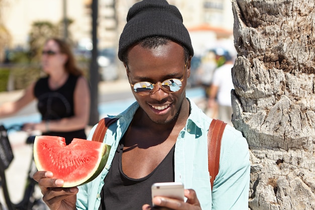 Gelukkige Afro-Amerikaanse toerist die verse, sappige watermeloen eet en 3G- of 4G-internetverbinding gebruikt op een mobiele telefoon terwijl hij ontspant op het strand, bij een palmboom staat en berichten van vrienden leest