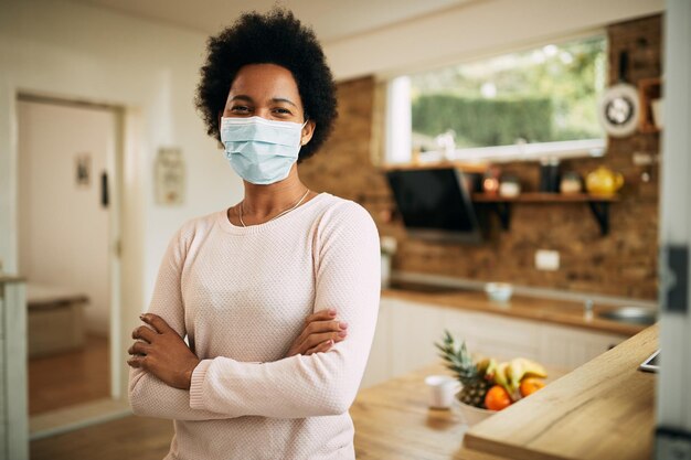 Gelukkige Afro-Amerikaanse vrouw die een gezichtsmasker draagt terwijl ze thuis met gekruiste armen staat