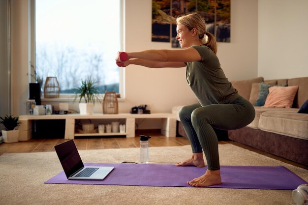 Gelukkige atleet die dumbbells gebruikt tijdens het oefenen van squats in de woonkamer