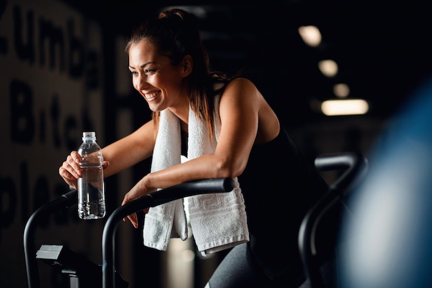 Gelukkige atletische vrouw die traint op een stationaire fiets en water drinkt in een sportschool