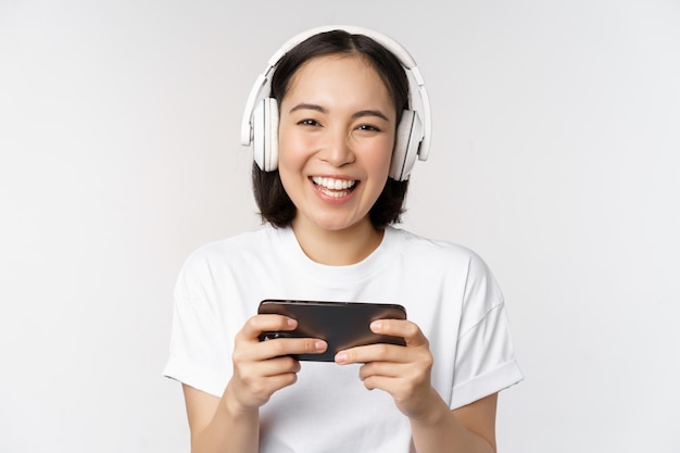 Gelukkige Aziatische meisjesgamer die op mobiele telefoon speelt die op smartphone kijkt die hoofdtelefoons draagt die zich over witte achtergrond bevinden