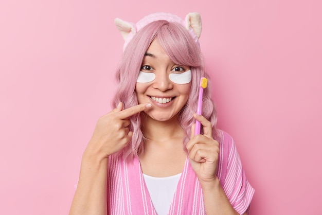 Gelukkige Aziatische vrouw met roze haar wijst naar een brede glimlach houdt tandenborstel ondergaat schoonheids- en hygiëneprocedures draagt casual kleding poses binnen tegen een roze achtergrond. Kijk naar mijn witte tanden