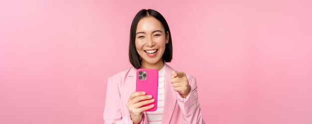 Gelukkige aziatische zakenvrouw die met de vinger naar je lacht en video opneemt die foto's maakt op een smartphone met een roze achtergrond van een mobiele telefoon