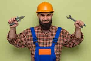 Gratis foto gelukkige bebaarde bouwman in bouwuniform en veiligheidshelm met moersleutel en tang die vrolijk glimlachend kijkt