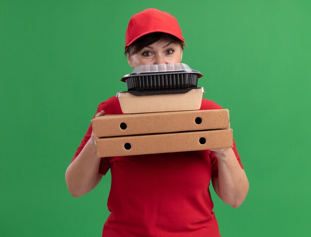 Gelukkige bezorgingsvrouw van middelbare leeftijd in rood uniform en pet die pizzadozen en voedselpakketten houden die voorzijde bekijken die zich over groene muur bevinden