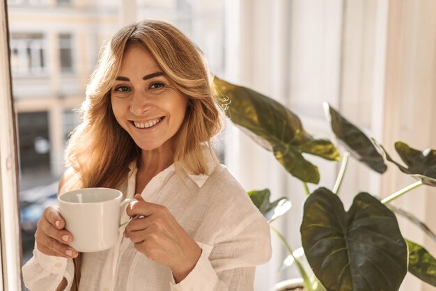 Gelukkige blanke volwassen vrouw die lacht en naar de camera kijkt terwijl ze 's ochtends koffie drinkt terwijl ze bij het raam staat Concept van rust en herstel