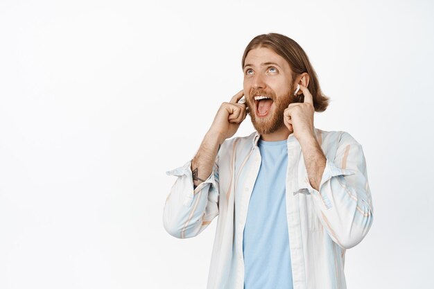 Gelukkige blonde man die muziek luistert op koptelefoons, draadloze oortelefoons, tevreden glimlacht en omhoog kijkt, meezingt met een lied, staande tegen een witte achtergrond.