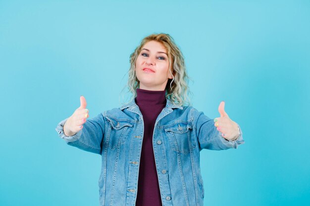 Gelukkige blonde vrouw kijkt naar de camera door handgebaren te tonen op een blauwe achtergrond