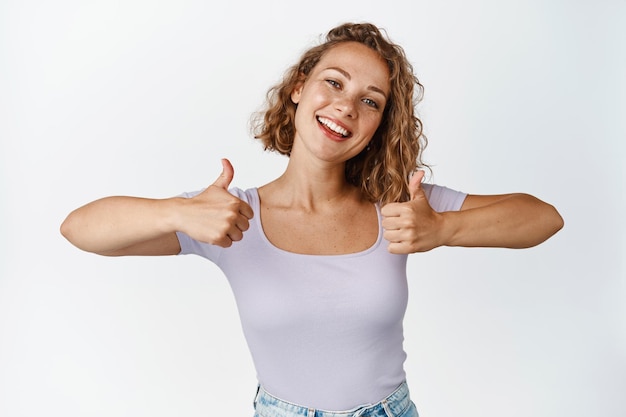 Gelukkige blonde vrouw toont duimen en glimlacht tevreden, prijst iets goeds, maakt complimenten, houdt van en keurt goed, staande op een witte achtergrond.