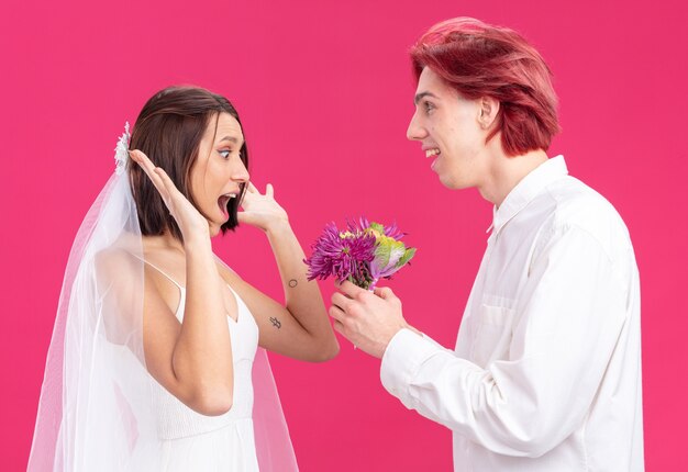 Gelukkige bruidspaar bruidegom en bruid gelukkige en vrolijke bruidegom die bloemen geeft voor zijn glimlachende en verraste bruid in trouwjurk die op roze staat