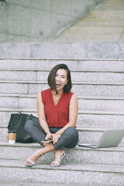 Gelukkige businsessvrouw laughong gezet op marmeren treden met dichtbij laptop, zak en meeneemkoffie