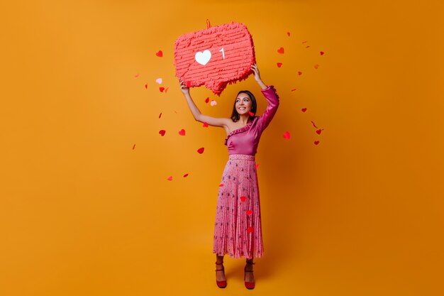 Gratis foto gelukkige, charmante vrouw van 23 jaar houdt een bord in de vorm van een like van instagram vast en poseert in volle groei op een oranje muur met confetti