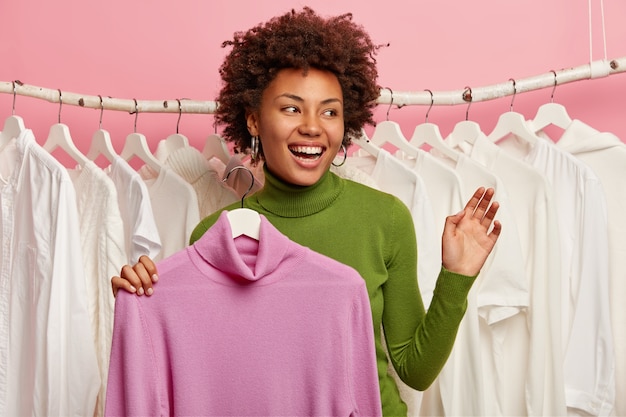 Gelukkige dame houdt nieuwe trui vast aan kleerhangers, houdt de hand omhoog, glimlacht breed, kijkt opzij, witte kleren hangen in rij achter.