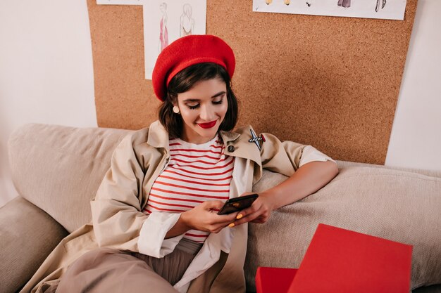 Gelukkige dame in heldere baret chatten in haar smartphone. Vrij mooie vrouw in moderne kleding zit op een bank en lacht.